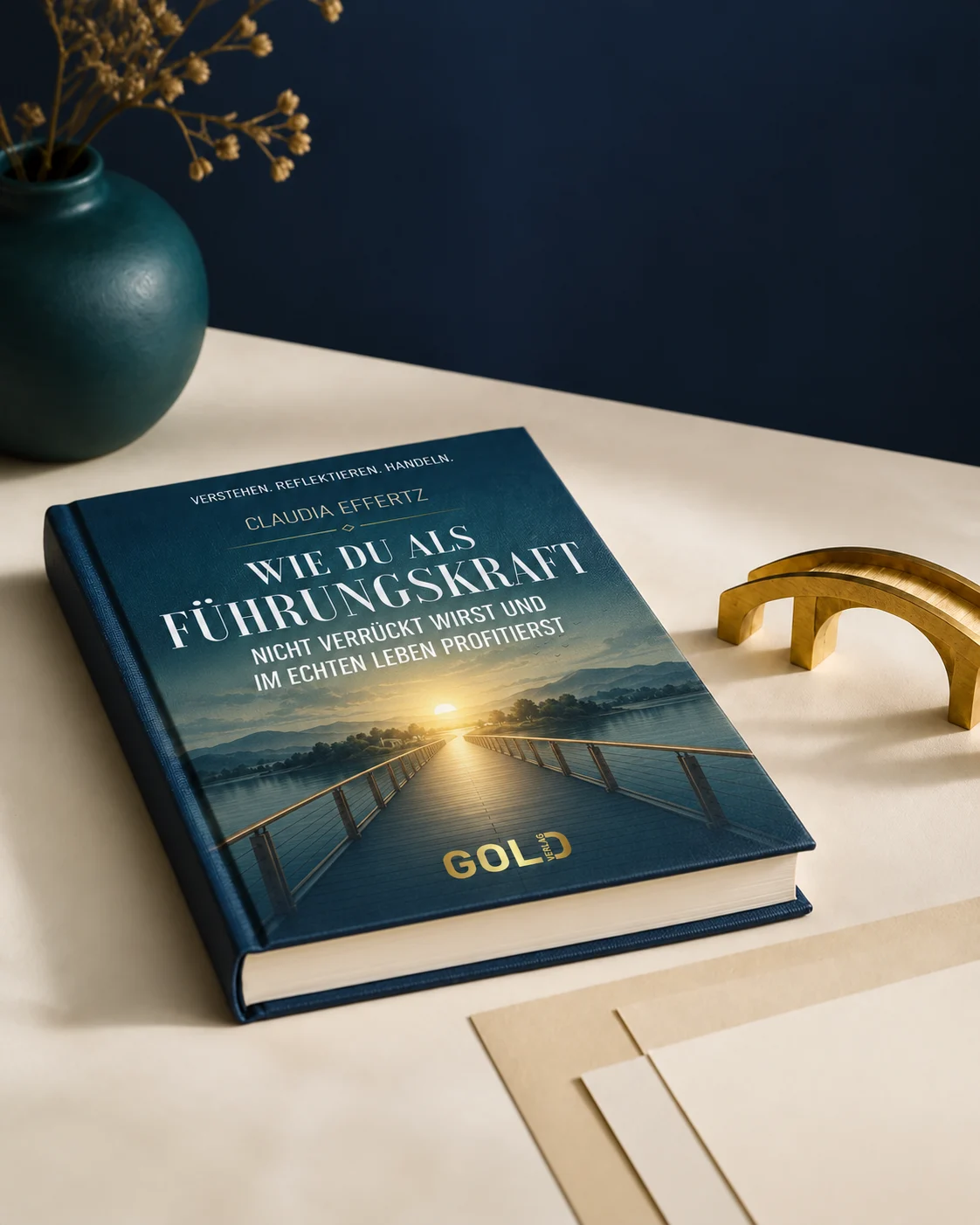 Claudias Buch auf einem hellen Tisch neben einer goldenen Brückenskulptur; auf dem Cover führt eine begehbare Brücke zum Sonnenaufgang